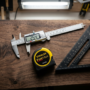 Professional measuring tools including digital calipers, a Stanley FatMax tape measure, and a black speed square on a walnut wood board.
