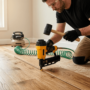 A photorealistic close-up of an experienced installer, wearing work gloves, kneeling on an oak floor, gripping a large pneumatic flooring nailer and mallet in mid-swing.