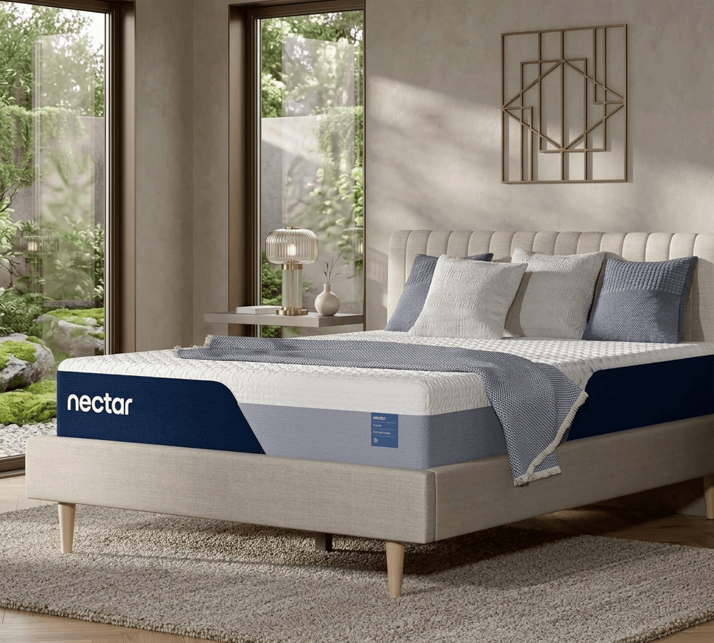 A detailed professionally styled photograph of a contemporary bedroom. The main subject is a Nectar brand queen-sized mattress (with visible 'nectar' logo) resting on a beige upholstered platform bed frame with warm wooden mid-century modern legs. The bed is dressed with two dark grey textured pillows, two light grey patterned pillows, and a textured grey throw blanket. To the left, a huge black-framed floor-to-ceiling window provides a stunning view of a beautifully landscaped garden featuring a large natural rock, various green trees, and flowering shrubs, with a distant city skyline under a clear sky. In the foreground, a modern, minimalist glass and brass table lamp sits on a small shelf near the window. The back wall is a neutral greige textured plaster, featuring a large, ornate geometric wire-frame sculpture above the headboard. To the right, a floating wood nightstand holds a white ceramic vase and a stack of books. A grey shag rug covers a portion of the light wood plank floor.