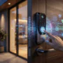 A close-up of a person using a biometric fingerprint scanner on a sleek modern smart lock with glowing digital security overlays showing connectivity and encryption icons.
