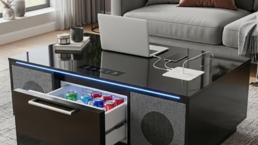 A modern living room featuring the Sobro Smart Coffee Table. The coffee table is sleek black with a glass top. One of the refrigerator drawers is slightly open, revealing cold drinks inside. On the other side, built-in Bluetooth speakers are integrated into the base. Subtle LED lighting glows beneath the table. The table is surrounded by a comfortable gray sectional sofa and is placed on a textured rug, creating a warm and contemporary atmosphere. A few items like a tablet and a small plant are on the tabletop.